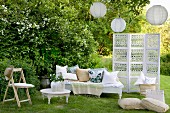 Romantic, white seating area in garden - bench with cushions and folding chair around side table next to screen below lanterns hanging in tree