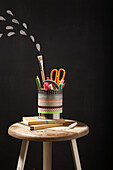 Office supplies and DIY pen holder made from tin can on wooden stool in front of blackboard wall