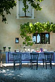 Long, set dining table with patterned tablecloth and blue wooden chairs outside restored property in southern France