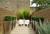 Gestylte Terrasse mit weissen Outdoormöbeln und formgeschnittenen Buchsbäumen in Pflanzgefässen vor Holzwand