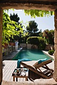 View though stone window frame of sun lounger on wooden deck next to Mediterranean pool
