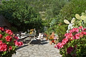Stühle und Tisch auf sonnigem Terrassenplatz mit Natursteinboden in blühendem mediterranem Garten