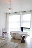 Helles Badezimmer mit freistehender Badewanne, raumhohem Fenster und Designerstuhl; im Vordergrund ein Waschtisch mit Mosaikfliesen