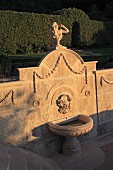In Abendsonne getauchte Steinmauer mit Skulptur und eingearbeiteten Verzierungen über Brunnen in Gartenanlage