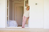 Older woman in pyjamas leaning on terrace door leading to bedroom