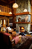 Gediegene, braune Ledersofagarnitur vor Kaminfeuer in offenem, rustikalem Wohnzimmer und Galerie aus Holz in warmem Braunton