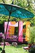 Modernes Tagesbett mit Baldachin aus pink und orangefarbenen Tüchern im sonnigen Garten; asiatischer Bambusschirm im Vordergrund