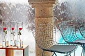 Wire chairs and roses in glass vases on side tables in front of stone column and waterfall sheeting into pool
