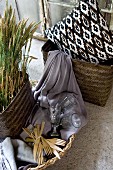 Basket of cereal ears next to wooden cutlery and glasses on lilac fabric and ethnic cushion in wicker basket