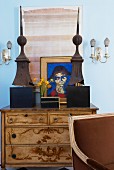 Partially visible chair in front of chest with marquetry and art objects in front of pictures of blue-painted wall