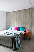 Doppelbett mit bunten Dekokissen vor Wand aus grauem Sichtbeton und Betonboden in modernem Schlafzimmer
