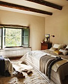 Comfortable bed with blankets and scatter cushions in front of open window in bedroom with wood-beamed ceiling