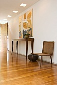 Modern painting above Colonial-style console table and 50s wooden chair in hallway with wide, swivelling wooden door