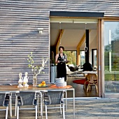 Essen auf sonnenbeschienener Terrasse - Blick durch offene Tür ins Wohnzimmer auf eine Frau mit Tablett und Gläsern in den Händen