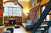 Loft-style interior with half-height shelving below arched window and grey leather sofa against brick wall