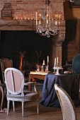 Lit candles in chandelier above small Rococo chair and table draped in table cloth in front of rustic fireplace in living room
