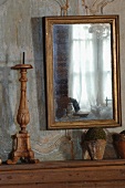 Gilt-framed mirror on faded wood-panelled wall and wooden candlestick next to vintage terracotta pots on console table