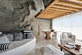 Curved bench with black and white patterned cushions against rock wall and white upholstered furniture in front of glass wall with transparent curtain