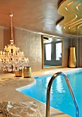 Chandelier with lit candles next to indoor swimming pool and suspended, curved golden ceiling panel