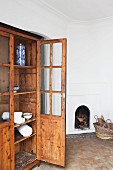 Old, glass-fronted cabinet with open door in interior with terracotta floor and open fireplace built into wall