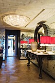 Console table with curved legs and table lamps with red lampshades against mirrored mosaic wall; elegant ceiling lamp on exposed concrete ceiling