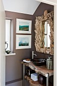 Rustikaler Waschtisch unter Badezimmerspiegel mit kunstvollem Rahmen aus Schwemmholz; an der Wand daneben zwei moderne Landschaftsaquarelle