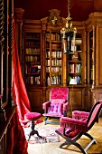 Antique chairs with pink velvet upholstery in front of antique fitted bookcase and red curtains at window in corner of grand library