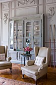 Antique, elegant reading chairs with white covers in front of glass-fronted display cabinet in painted, wood-panelled wall with stucco elements