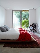 Simple double bed with red blanket on Oriental rug in front of sliding French windows with view into garden
