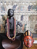 Afrikanische Holzstatuette mit daran hängenden Halsketten auf poliertem Holzobjekt und Holzmaske vor chinesischem Wandbild