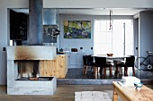 Concrete open fireplace in open-plan interior with dining table and chairs next to industrial window on black-tiled platform