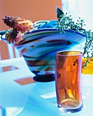 Cylindrical glass in front of glass vase of red hot pokers and stalks of buds on glass table top
