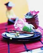 An Asian place setting with duckweed and chopsticks on a red bamboo table runner