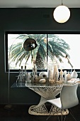 Collection of white vases and sculptures on modern glass table next to arc lamp and vintage chair in front of window with view of palm tree