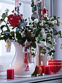 Baubles on branches of snowberry bush in white vintage pitcher and lit candles on window sill