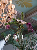 Silberfarbene Vogelfiguren auf Zweige geklipst unter leuchtendem Kronleuchter