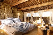 Double bed and tables made of tree trunks on sand floor in barn-like interior with rough stone walls and gathered curtains at window