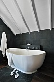 Freestanding bathtub in a modern bathroom with slate tiles on the wall and floor under a wood ceiling painted white