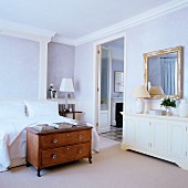 Antique wooden cabinet at foot of bed and white sideboard against wall next to open door in lilac bedroom with stucco ceiling frieze