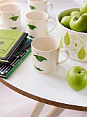 Tassen mit Apfelmotiv neben Schale mit grünen Äpfeln auf weißem Tisch