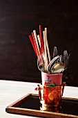 A mix of European and Asian eating implements - chopsticks and cutlery in stacked cups on a wooden tray