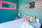 Colourful scatter cushions on bed below pink-framed mirror and artworks with mottoes in English on turquoise wall