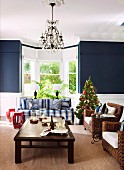 Small Christmas tree between blue and white striped sofa and wicker armchair in front of living room bay window flanked by blue-painted walls