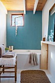 Simple bathroom with bathtub against light blue wall and small window; towels on chair and sisal runner in foreground