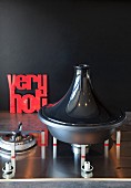 Elegant tagine pot on gas hob and red lettering ornament leaning on black wall