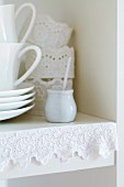 Mit weißem Spitzenband verzierte Kante eines Küchenregals; Serviettenringe aus gestärkter Spitzenbordüre im Hintergrund