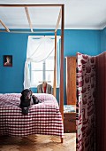 Bett mit einfachem Baldachingestell und Hund auf karierter Tagesdecke im himmelblau getönten Schlafzimmer