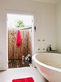 Moderne Badewanne in weißem Badezimmer, Blick durch offene Tür auf mit Bambusstäben eingezäunte Terrasse mit aufgehängtem, rotem Handtuch