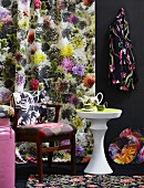 Tea set on side table and upholstered chair in front of floral panel curtain and dark background