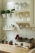 Cups hanging from hooks below shelves mounted on tiled wall above antique kitchen scales on wooden worksurface
